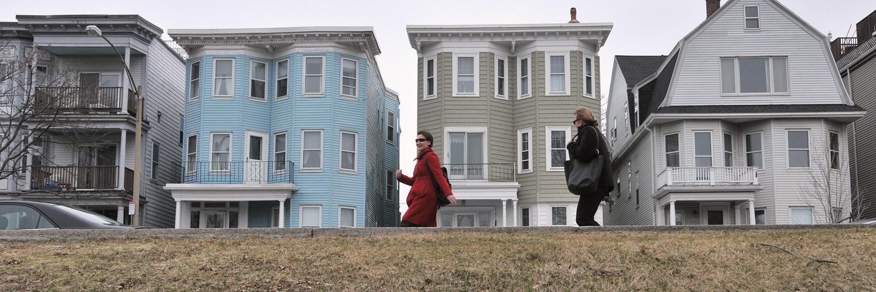 Two people walk past triple decker houses in Dorchester.